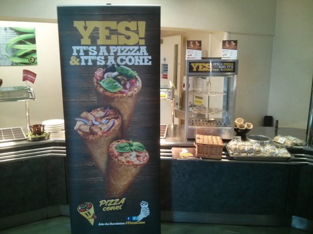 Pizza Cones banner reading 'Yes! It's a pizza & it's a cone' beside a display cabinet at a food counter