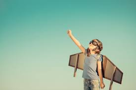 Child in a pilot's cap and goggles wearing cardboard wings, one arm raised to the sky