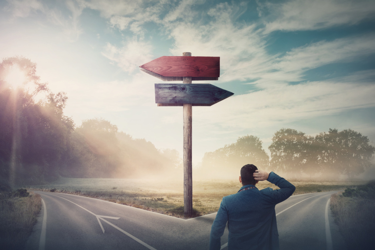 A man scratching his head at a fork in the road beneath a signpost pointing two ways
